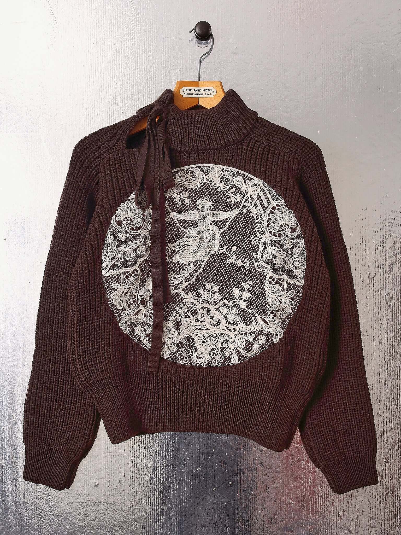 A flat shot of the 'Circle' sweater on a hanger showing the asymmetric tie neck line and the circular embroidered knit inspired by the famous painting 'The Swing'.