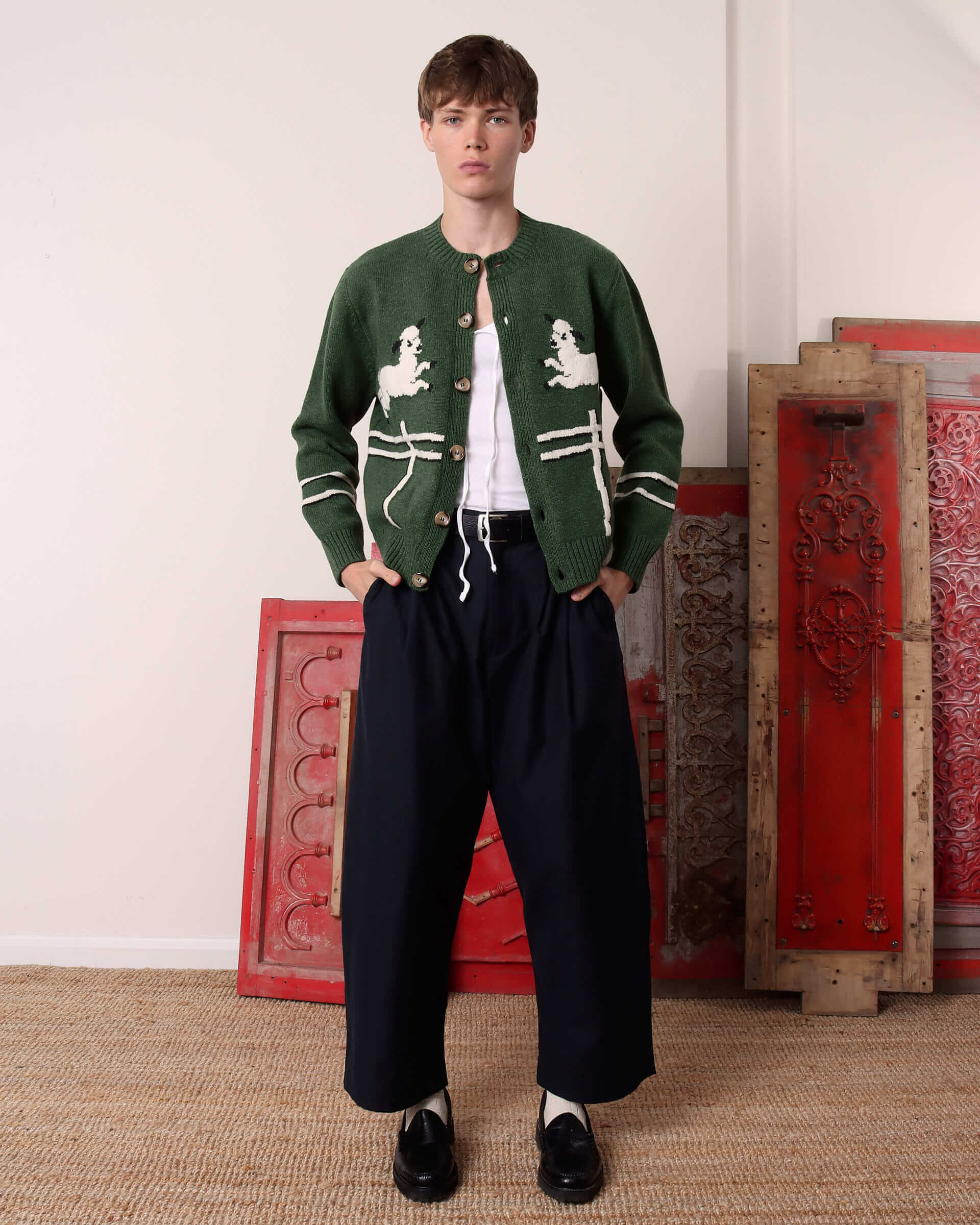 A model wears the 'Cyril' Lamb Motif Cardigan in green unbuttoned with a white vest underneath and with a pair of black trousers.