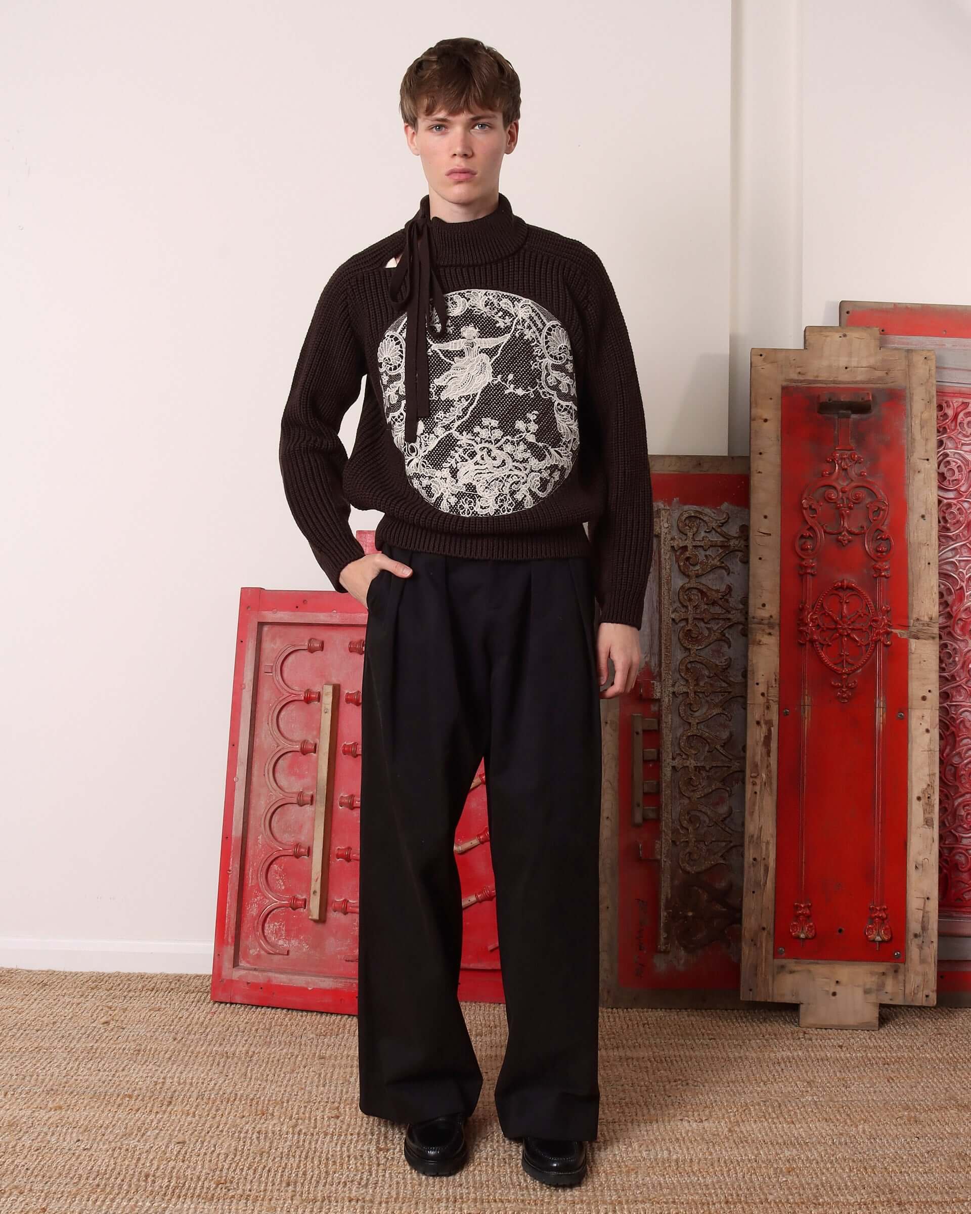 A Model wears the 'Circle' Sweater in brown marino wool, with matching brown pleated trousers.