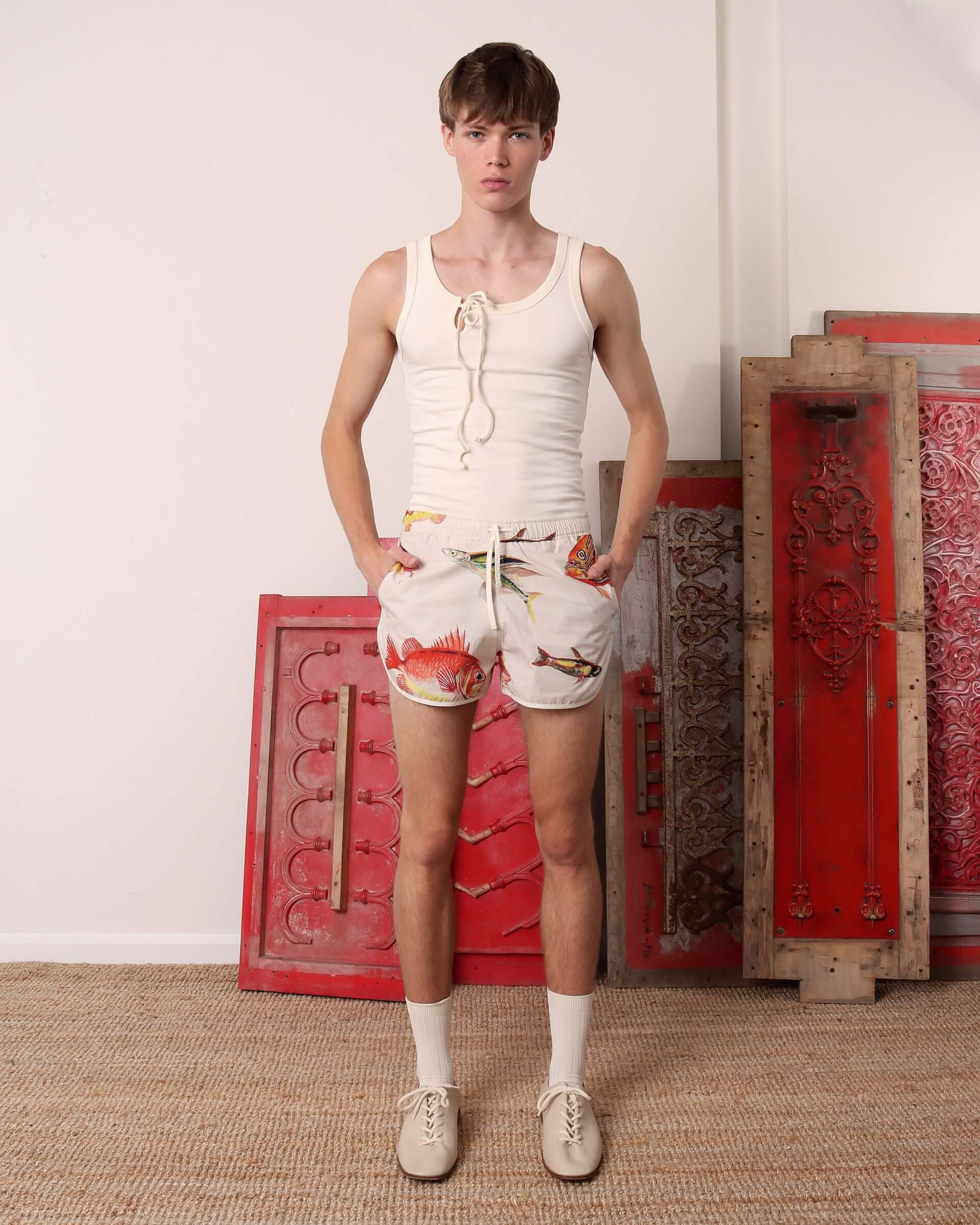 A model wears the ecru 'Cochrane' Vest styled with a fish print drawstring short with his hands in the pockets.