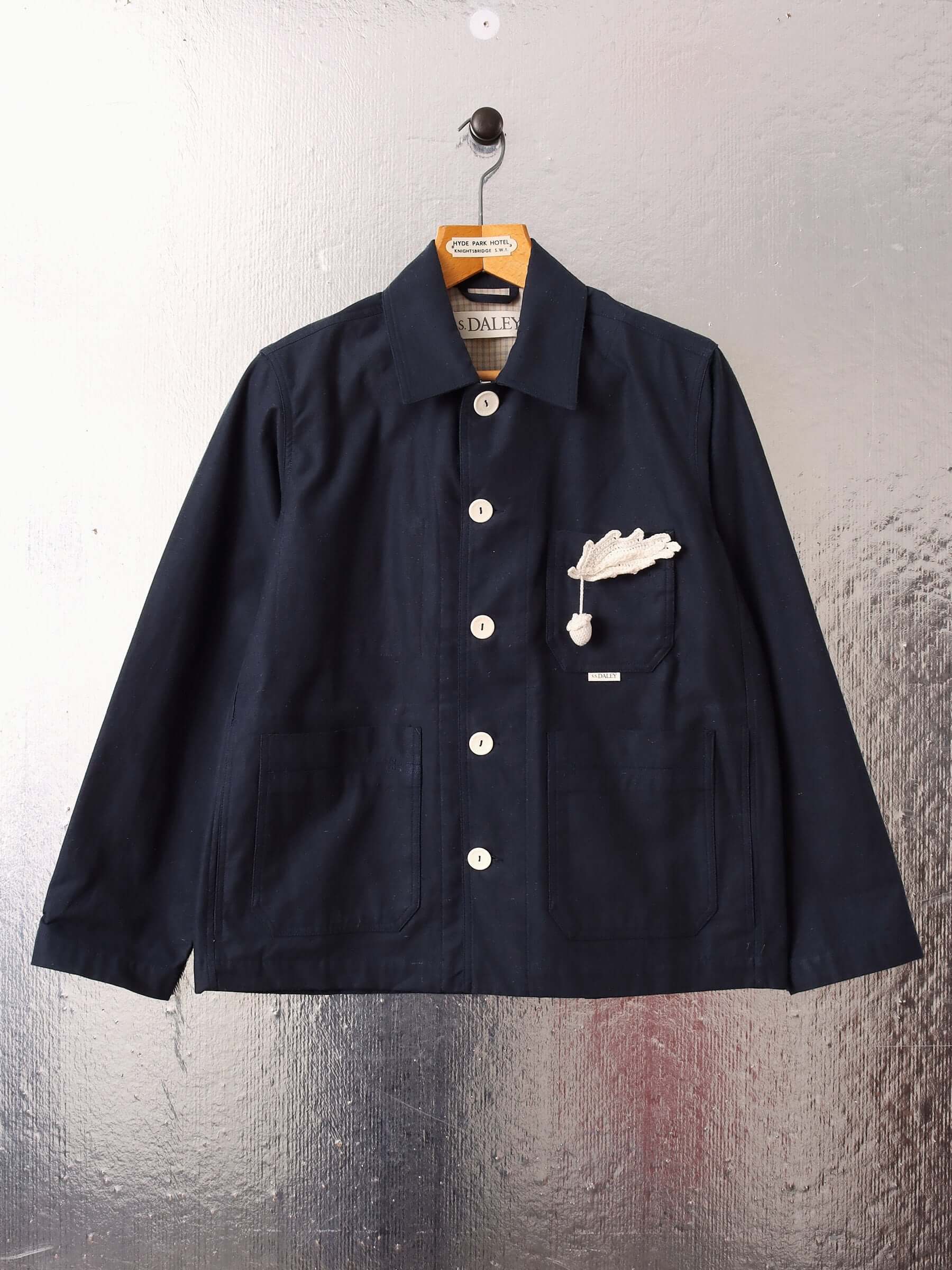 A flat shot of the 'Clarence' Chore Jacket in navy showing the front five white buttons buttoned up to the collar and a white hand-crocheted acorn pin.