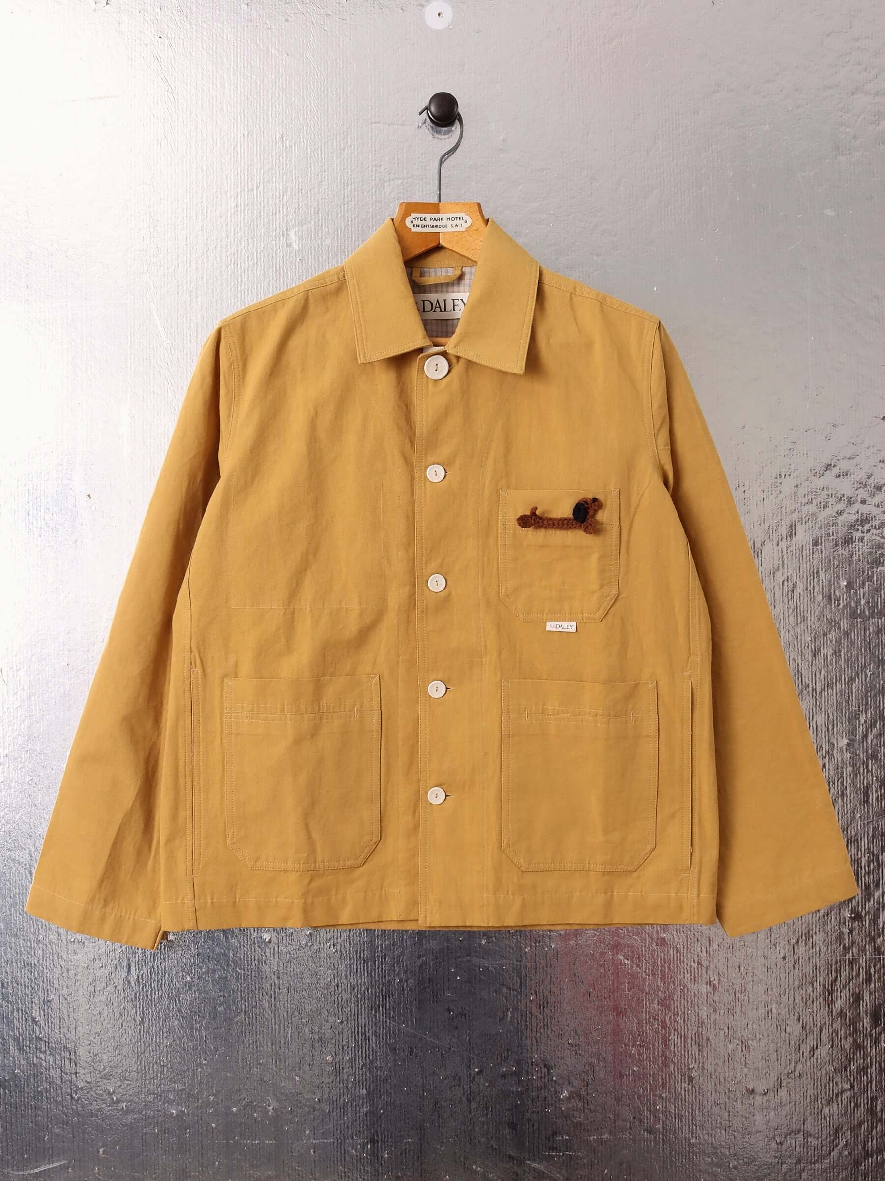 A flat image of the 'Clarence' Chore Jacket in mustard, with the ecru buttons buttoned up to the collar and the removable Dachshund pin in the front chest pocket.