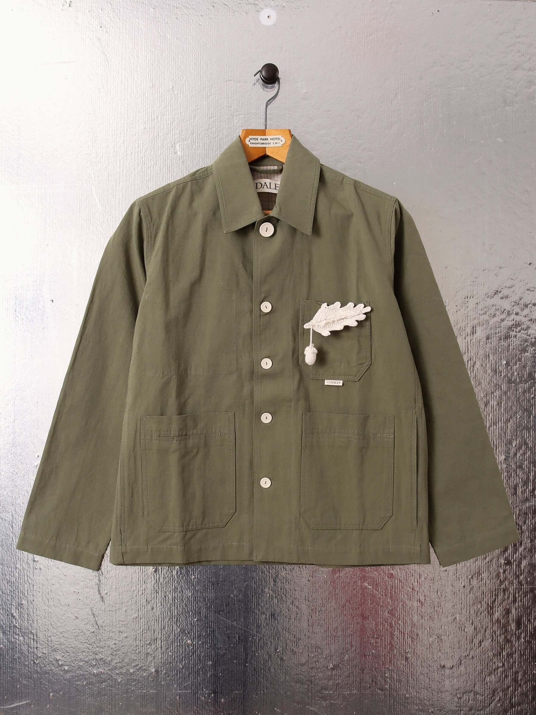 A flat shot of the front of the green 'Clarence' Chore Jacket with its ecru buttons done up to the collar showing the two front pockets and third chest pocket with a detachable crocheted acorn pin.