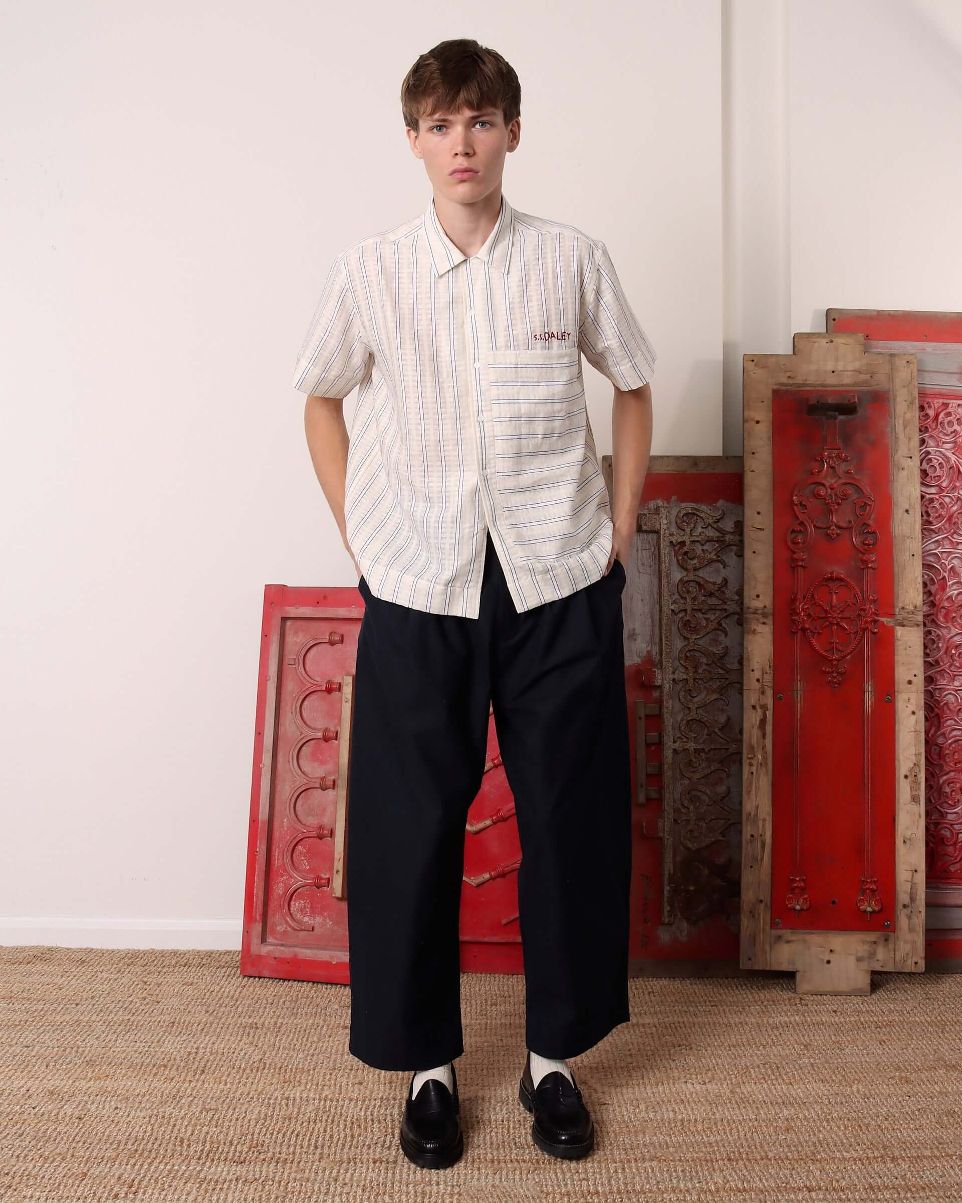 A model wears the striped 'Turing' Shirt with short sleeves buttoned up to the top and styled with black pleated trousers.