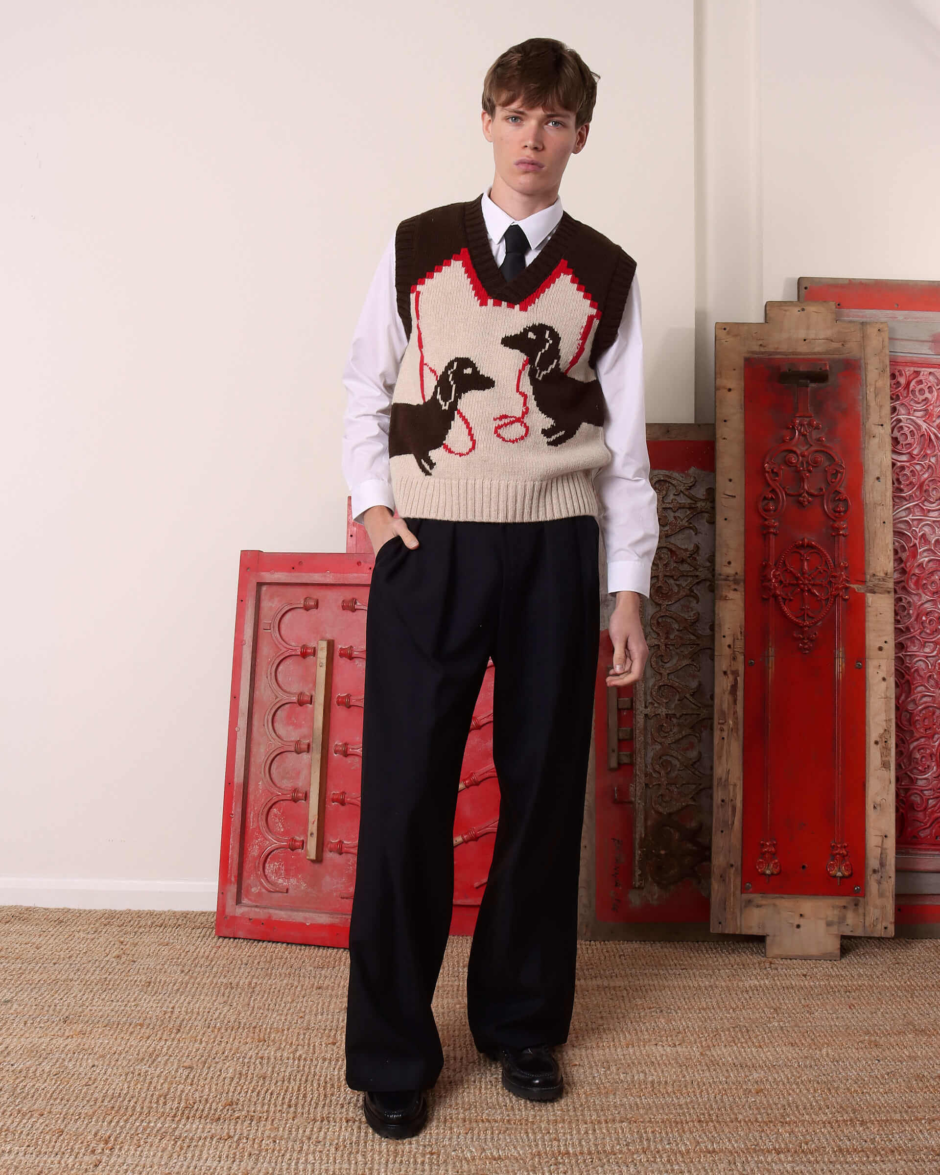 A model wears the 'Harold' Sweater Vest in brown and ecru showing the brown dachshund dog motifs and red leash designs.