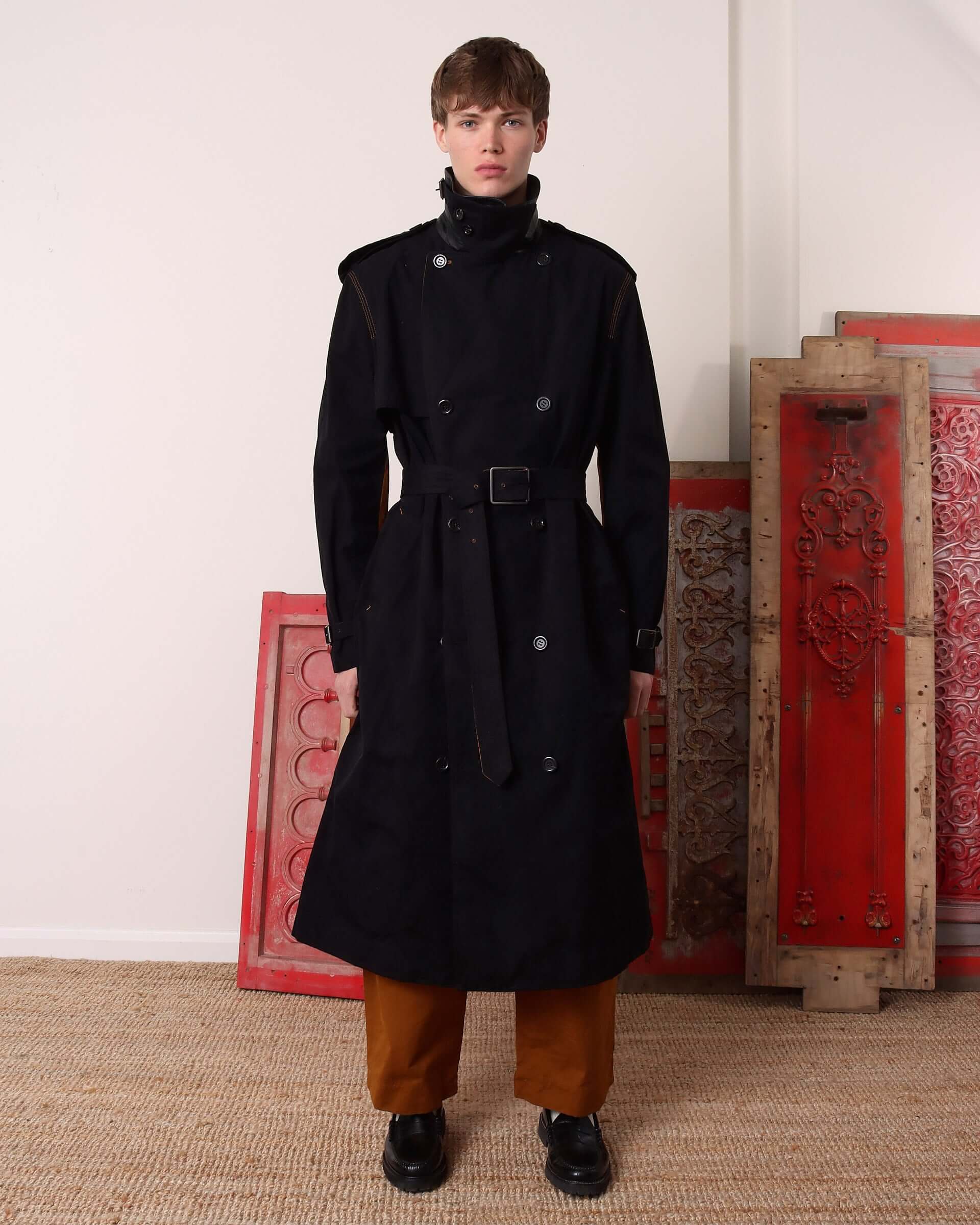 Model wearing a black trench coat with brown trousers.