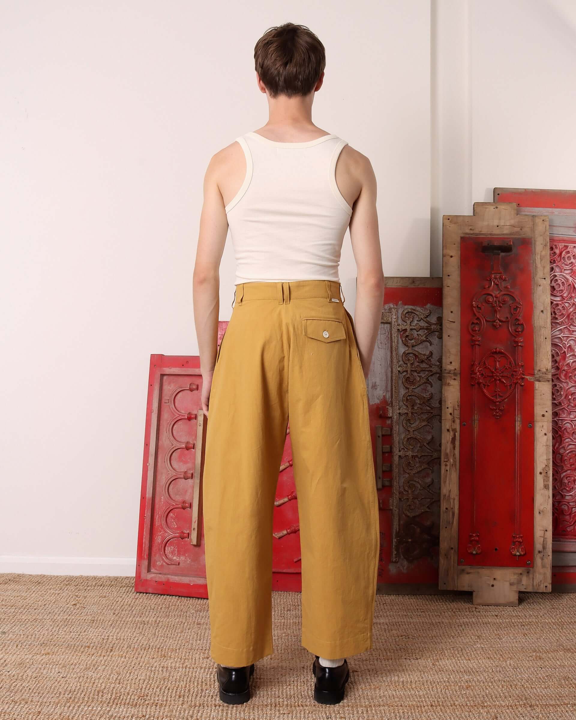 A model shows the back of the 'Clarence' Trousers in mustard, showing the right side back pocket with a white button closure.
