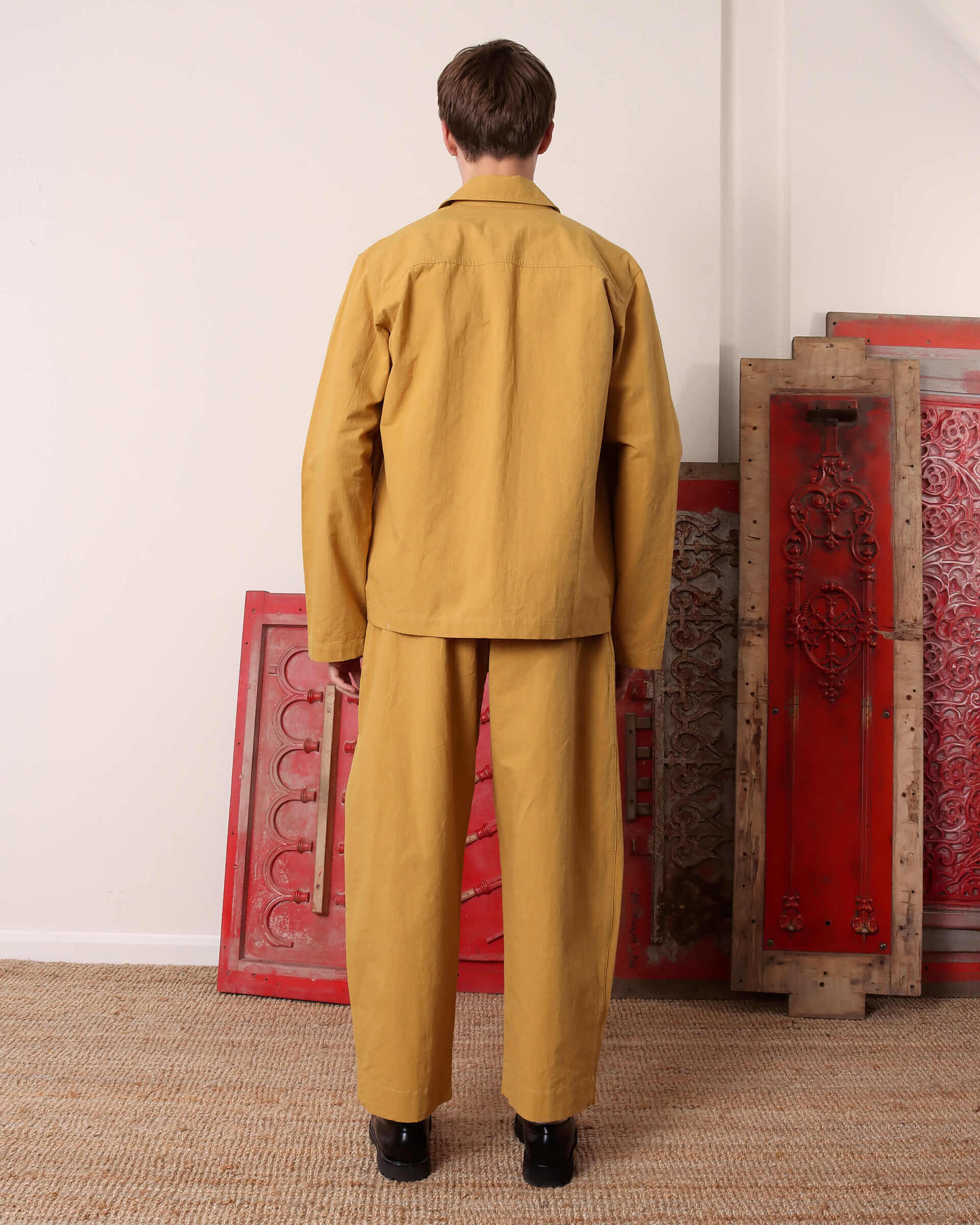 A model shows the back of the 'Clarence' Chore Jacket in mustard and matching 'Clarence' Trousers in mustard.