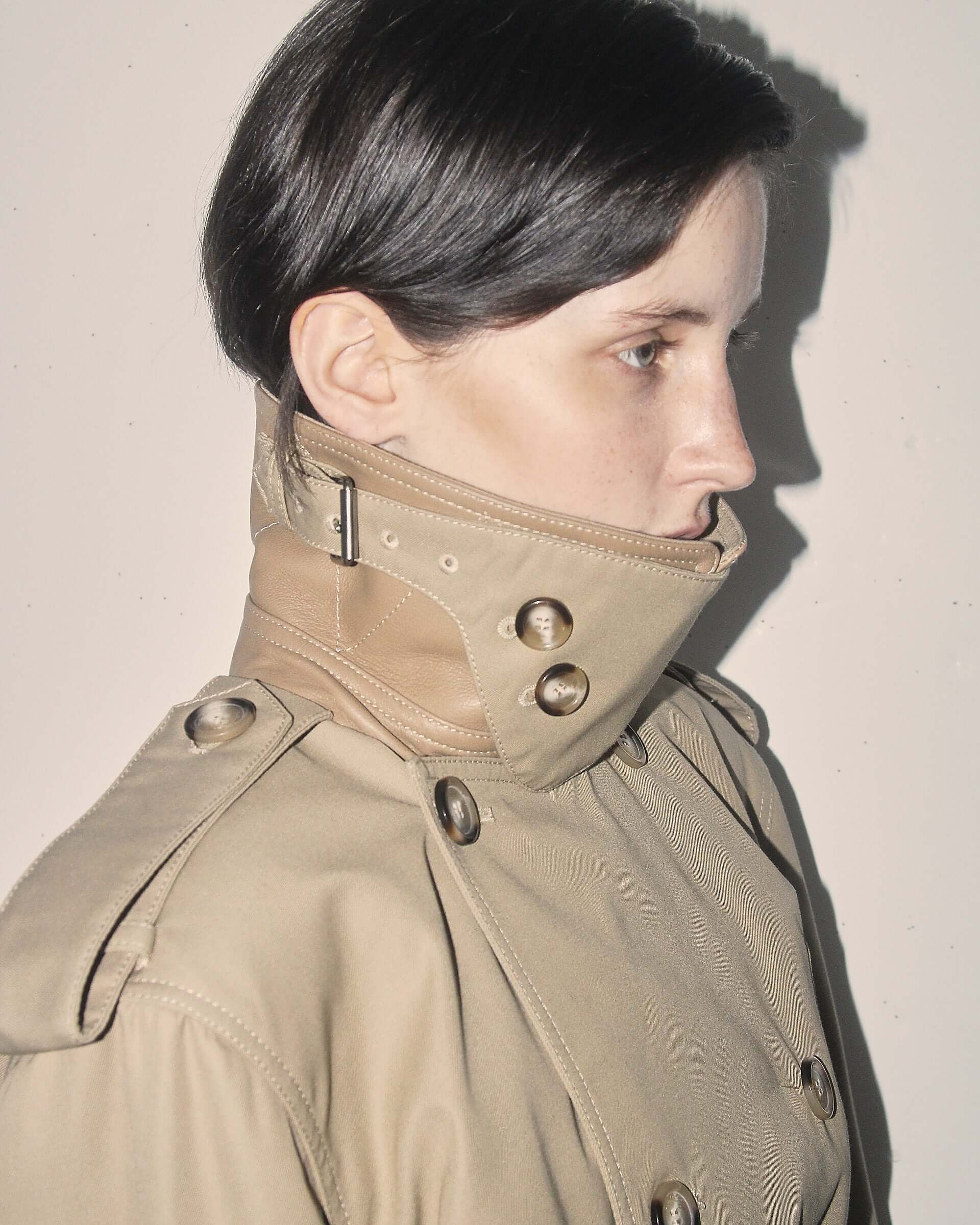 A detail shot of a model wearing the 'Eliza' Peplum Trench with the adjustable buckled neck closure with button details fastened and an epaulette design on the shoulder.