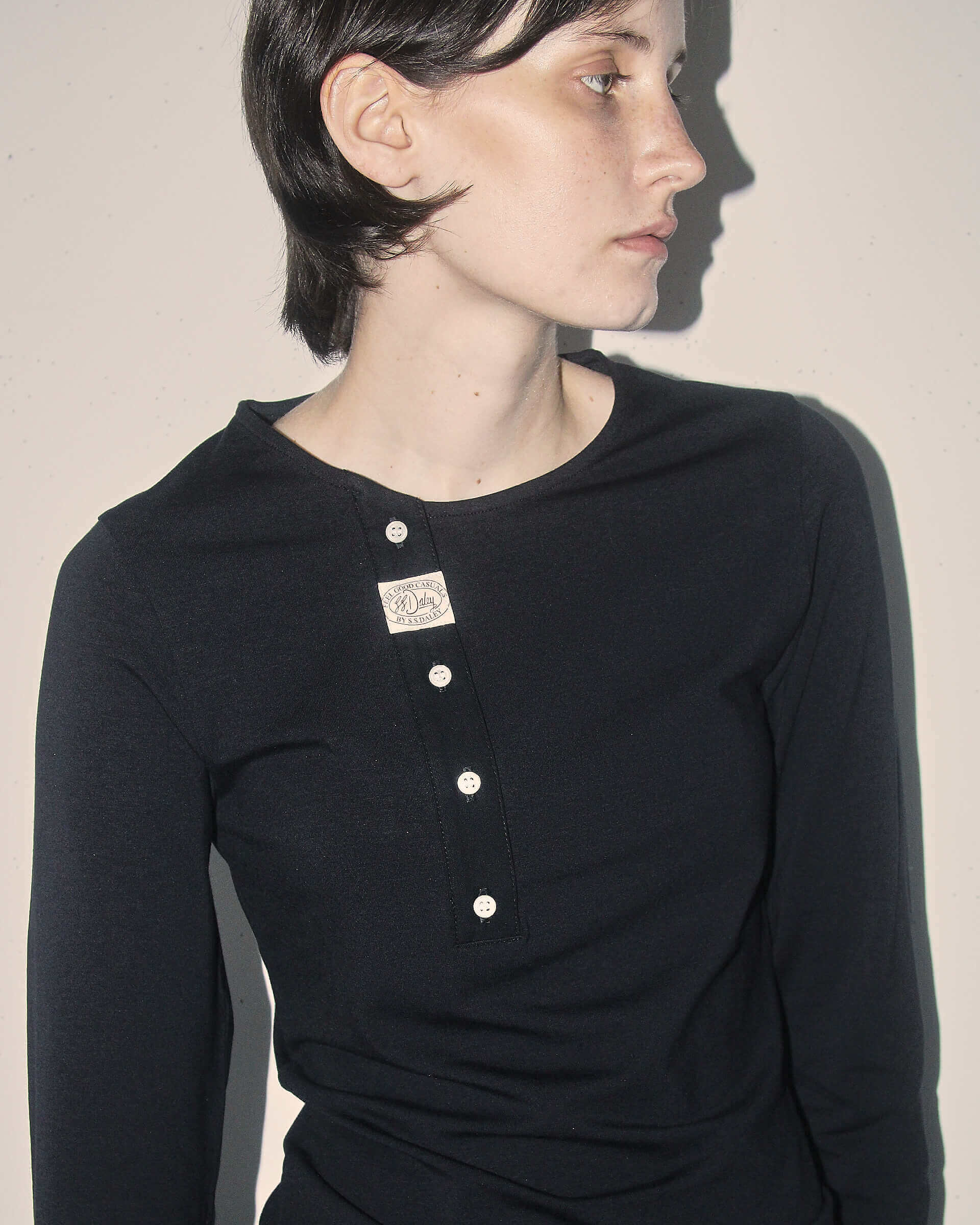 A detail shot of a model wearing the black 'Aker' Henley, showing its asymmetric placket design, ecru buttons, and S.S.DALEY Feel Good Casuals label.