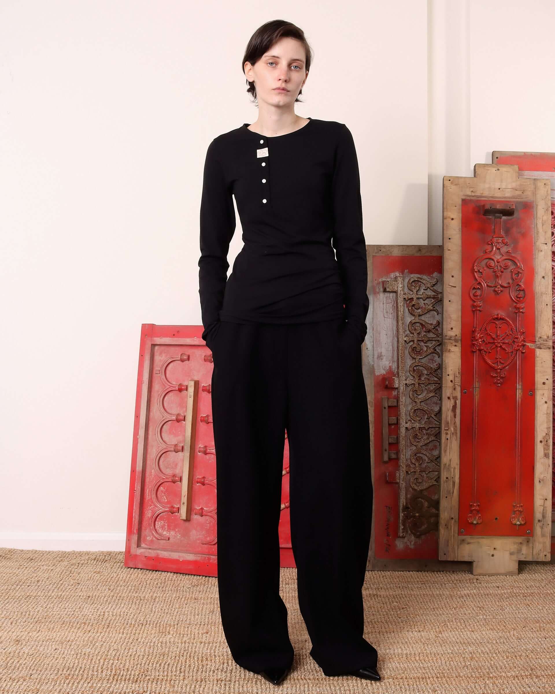 A model wears the black 'Aker' Henley with matching black trousers with her hand in the pockets.