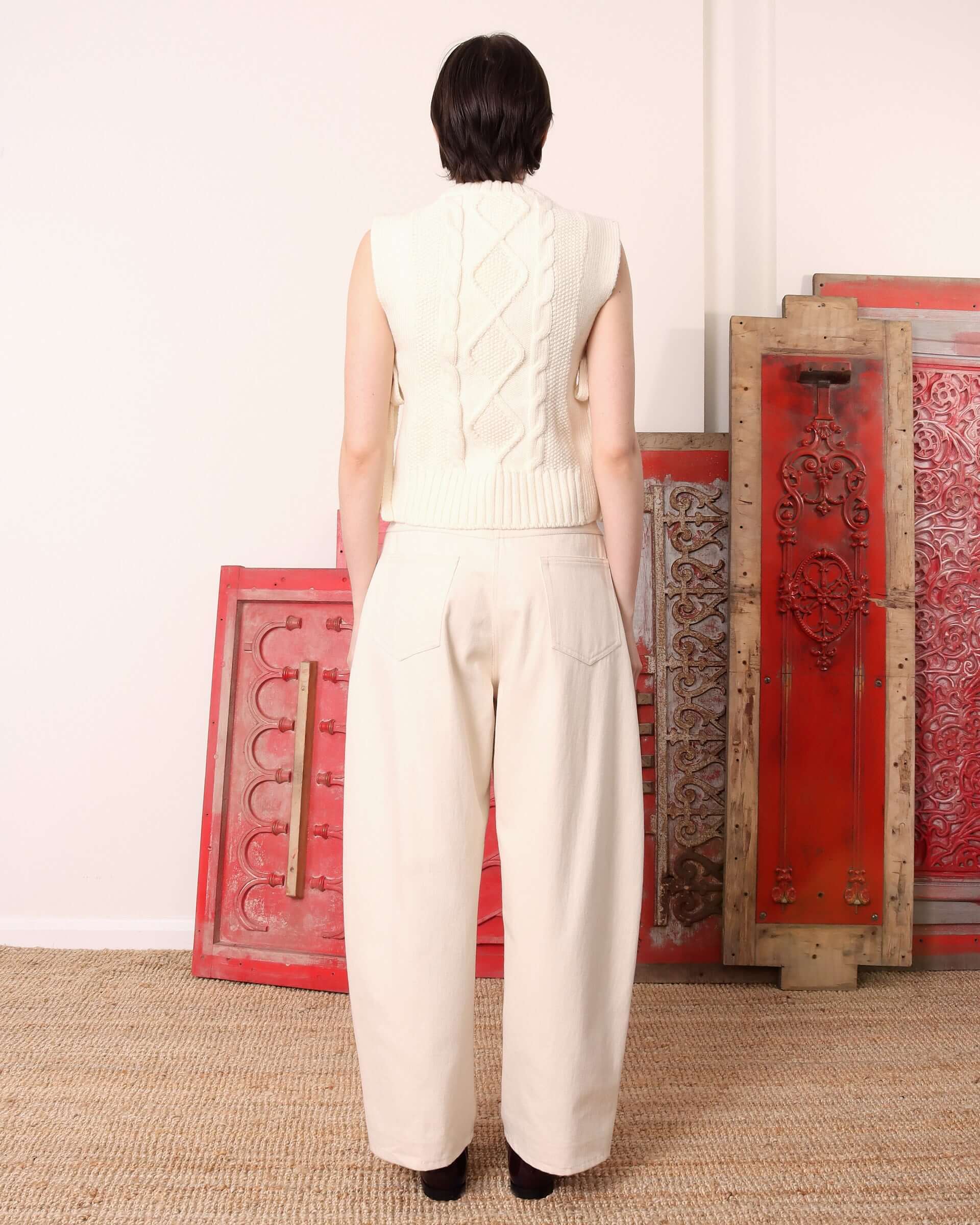 The model shows the back of the ecru 'Blandine' vest which shows the back cable knit design and ribbed hem.