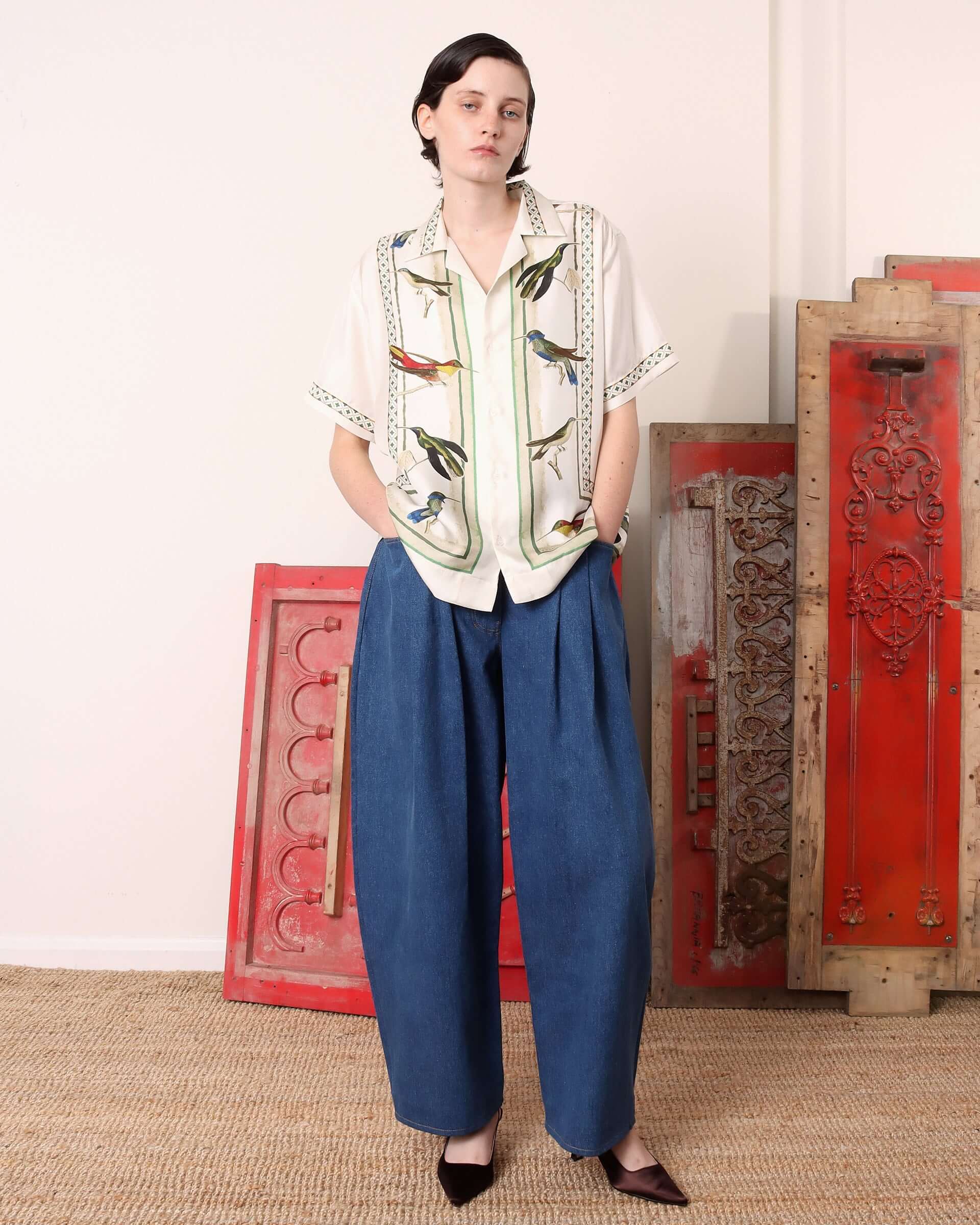 Model wearing the short sleeve 'Birdwatcher shirt, featuring a print of colourful hummingbirds and intricate  green weave and stripe designs.