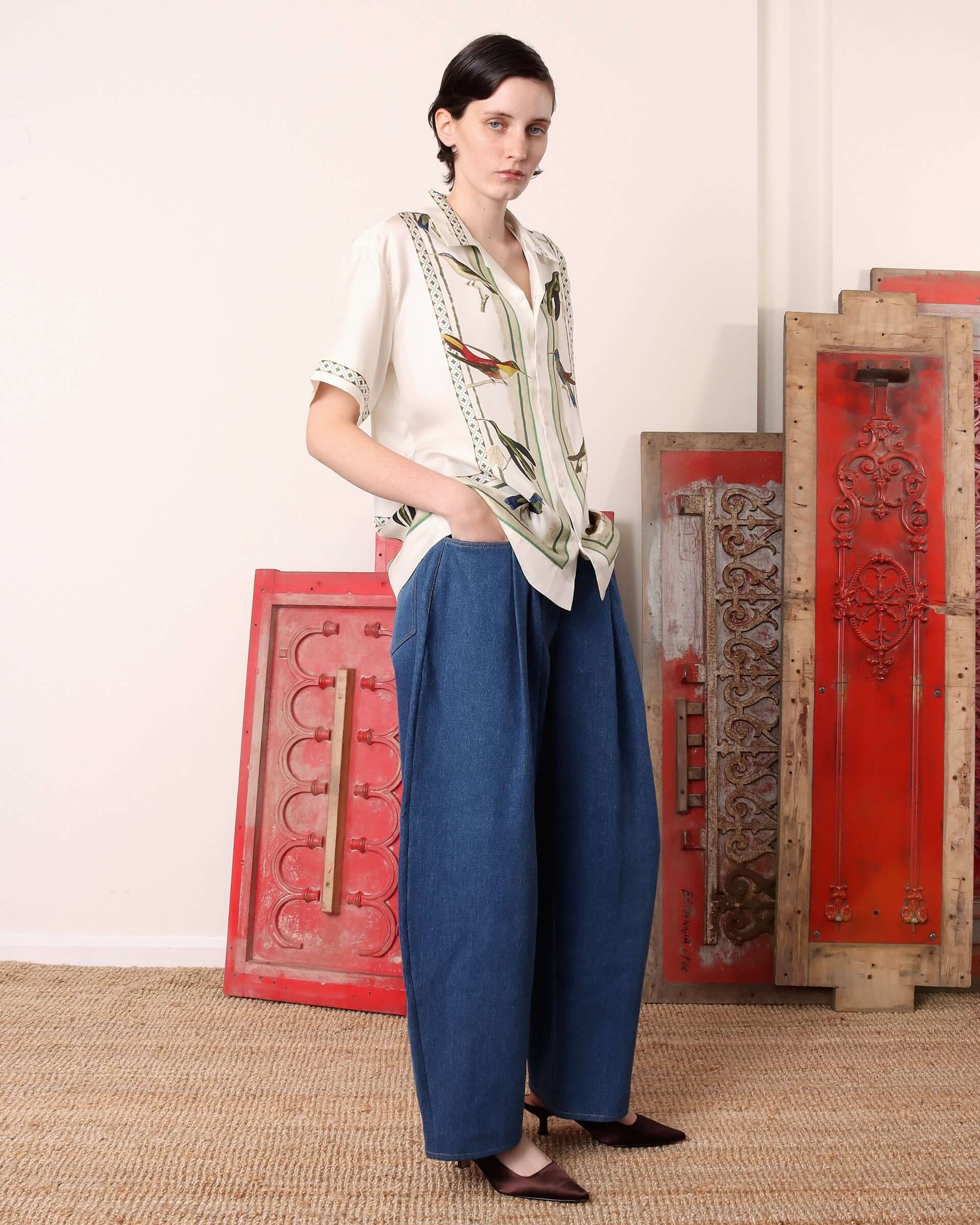 A model wears the 'Birdwatcher' shirt and has her hands tucked into the pockets of the denim trousers.