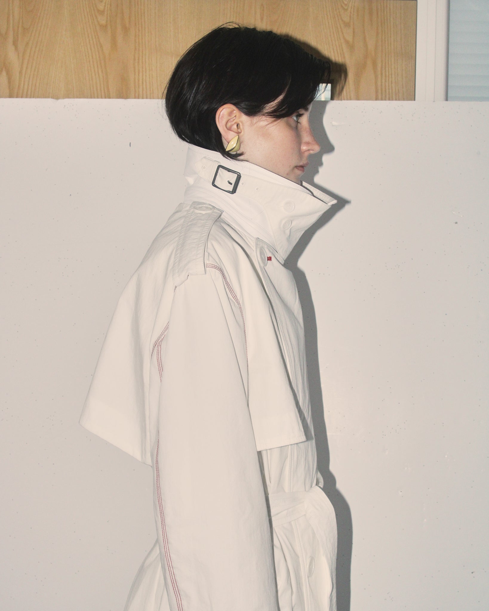 A detail shot of a model showing the side profile of the 'Sophia' Trench in white, showing the throat latch closure and collar up. 