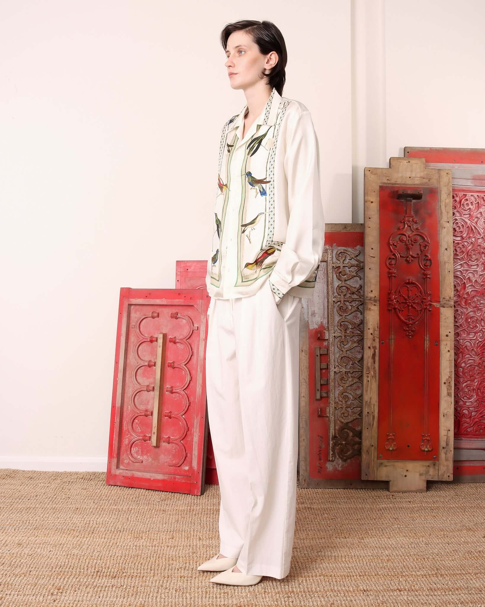 Model wearing the 'Birdwatcher' shirt buttoned up, with her hands in the pockets of the matching white trousers. 