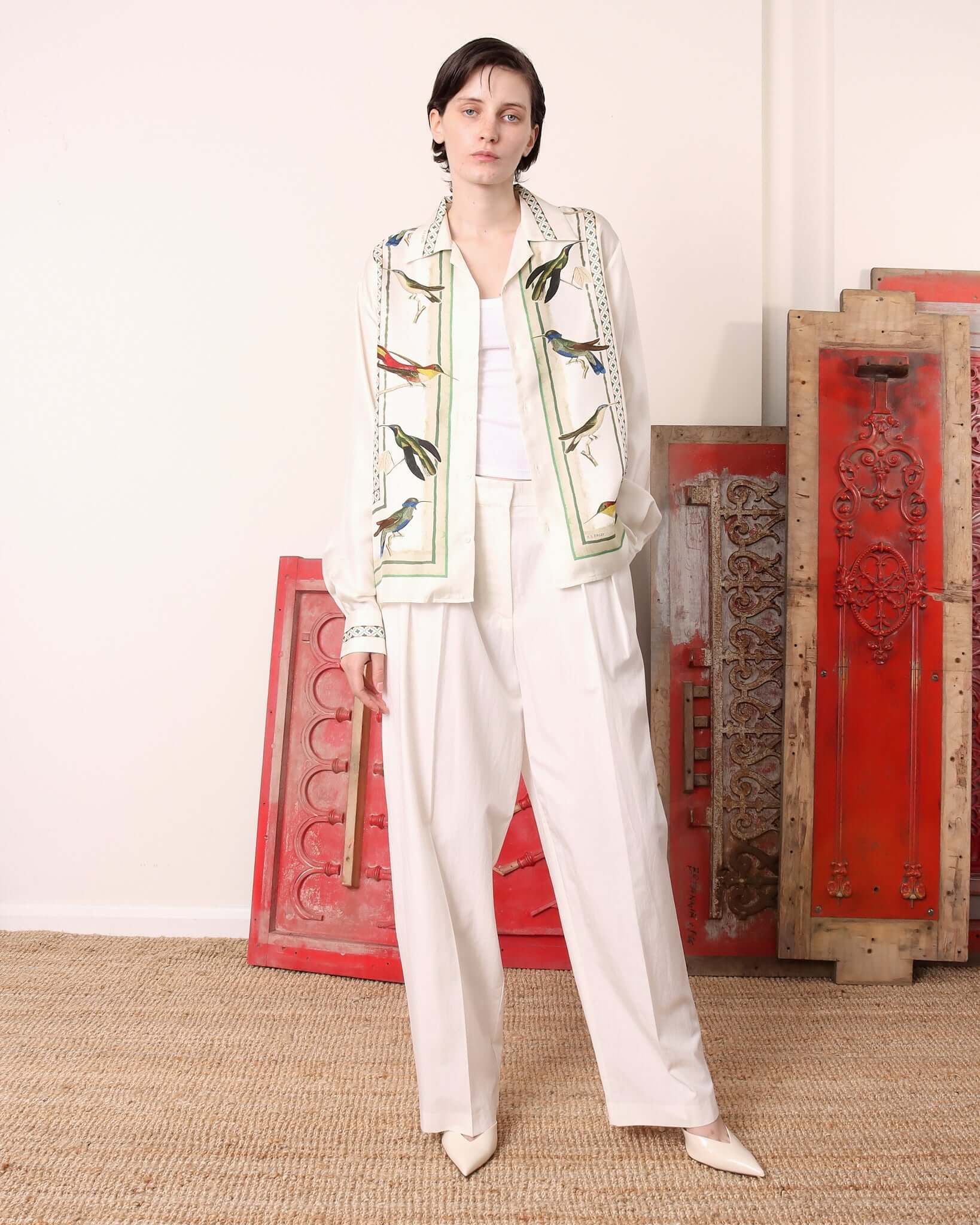 Model wearing the 'Birdwatcher' shirt with long sleeves, featuring an intricate design with green lines and weaves on a light ecru background with multiple colourful, realistic birds. 