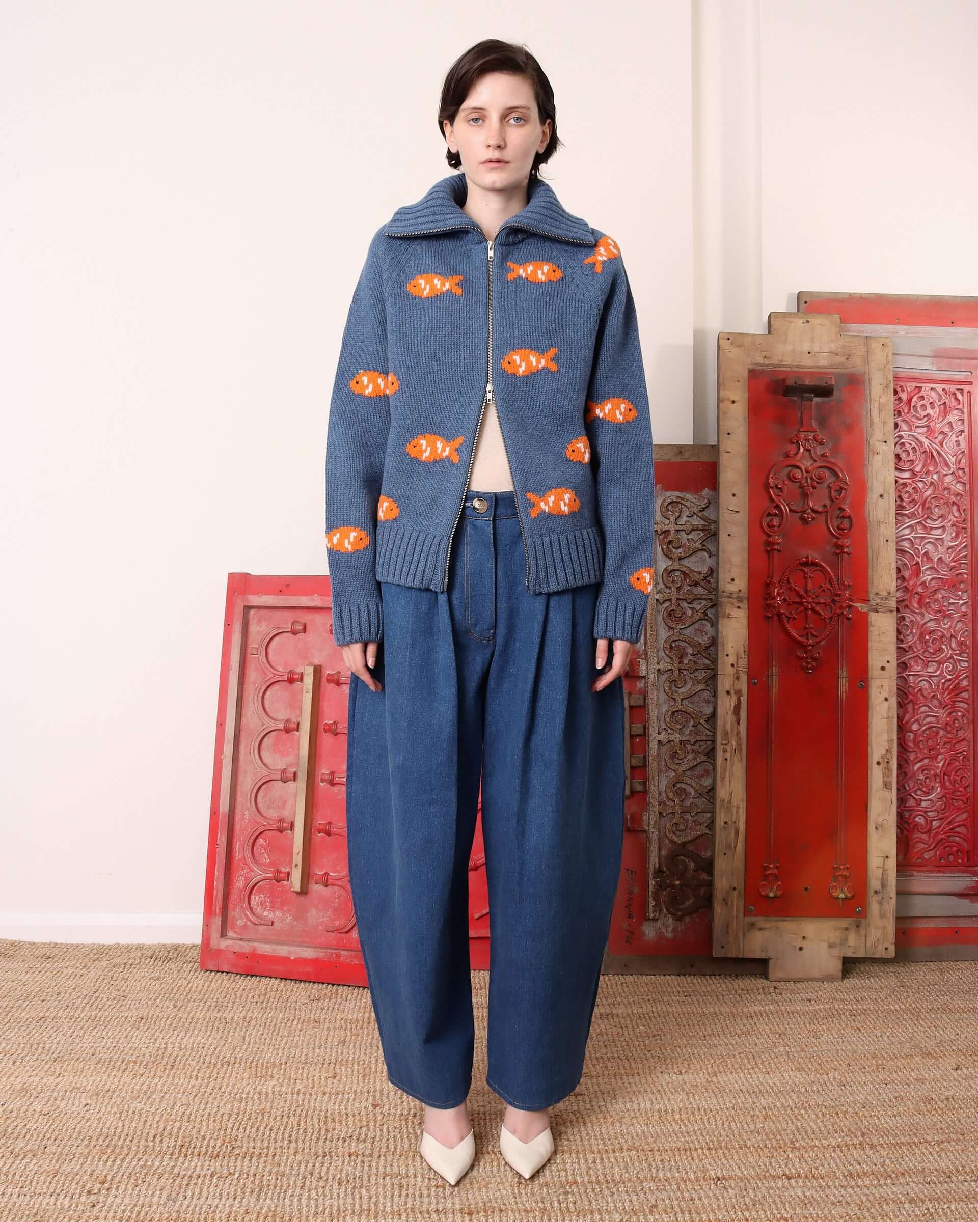 A model wears the 'Archie' knit zip-up cardigan in ocean blue with an intarsia goldfish design styled with pleated denim jeans.