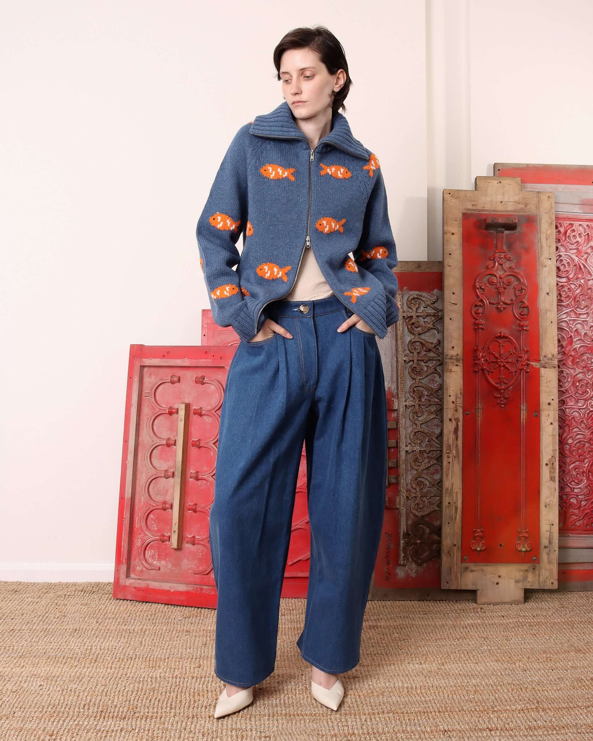 Model wearing the 'Archie' Knit blue zip-up sweater with a ribbed fold-over collar and orange goldfish intarsia designs. 