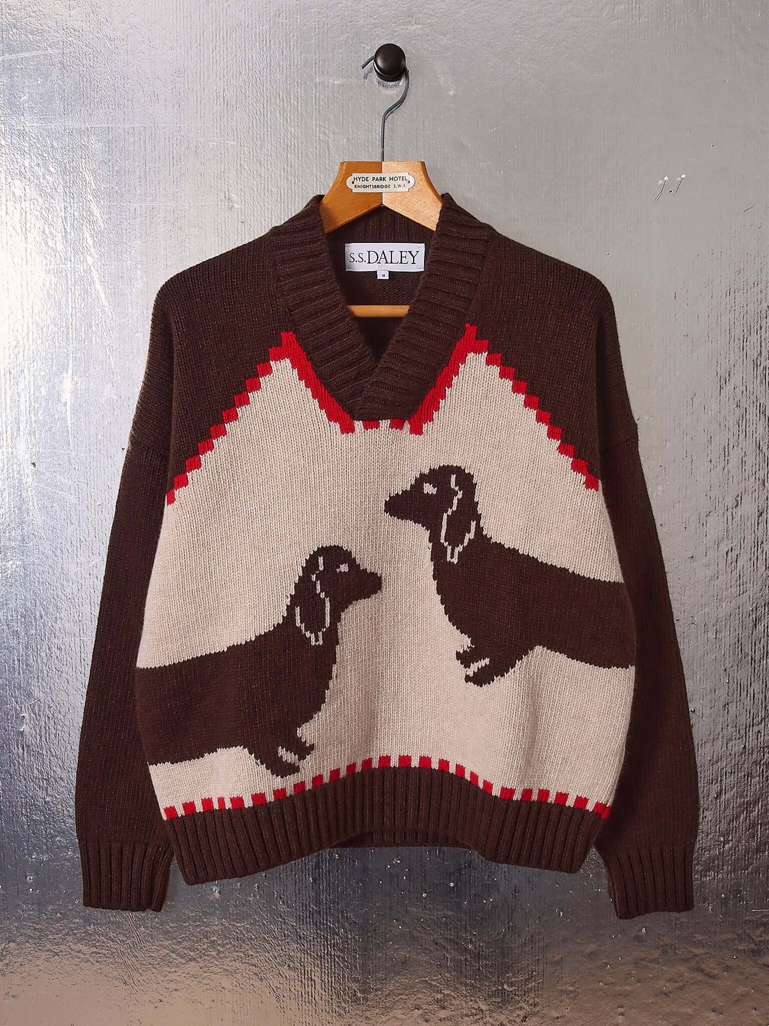 A flat shot of the 'Harold' V-Neck Sweater Vest in brown and ecru on a hanger showing the brown, ecru, and red colourway and two sweet dachshund motifs.