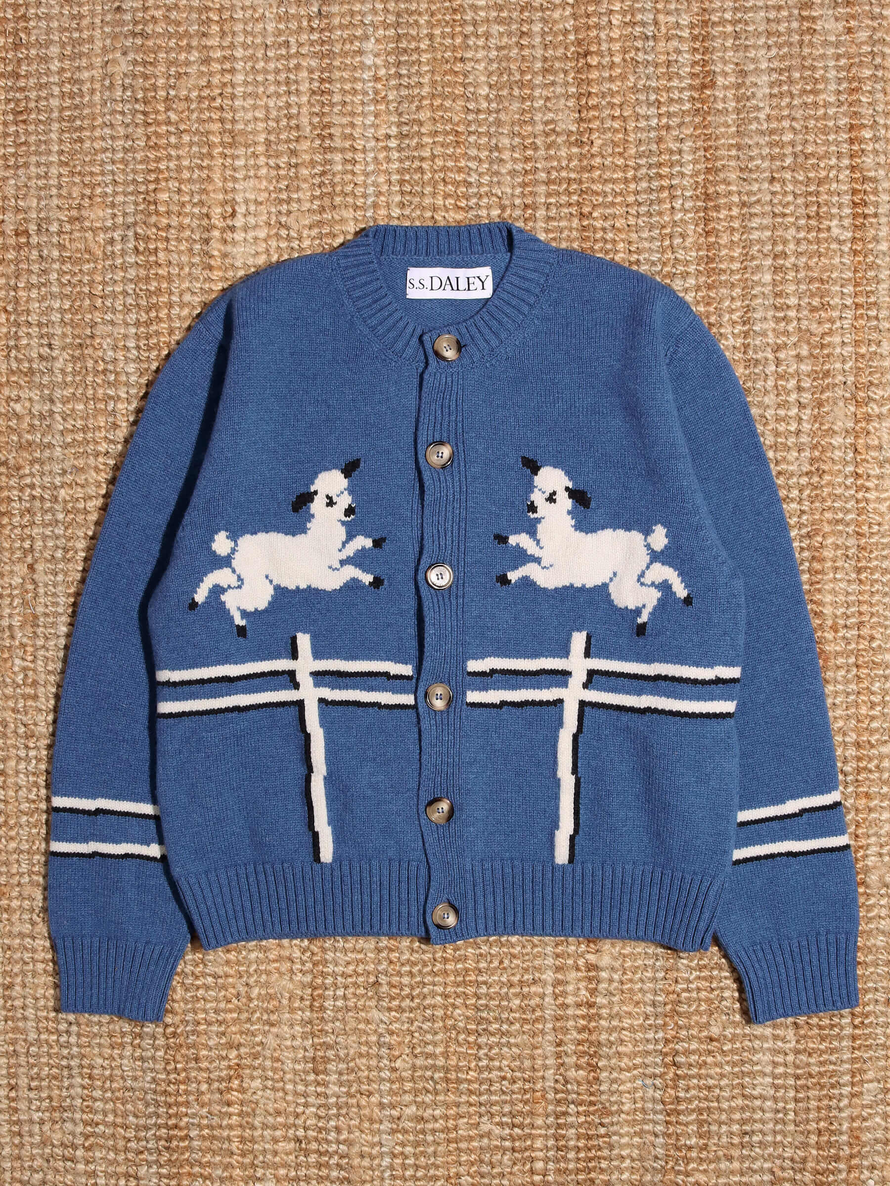 A flat shot of the 'Cyril' Lamb Motif Cardigan in blue showing its two lamb and fence motifs.