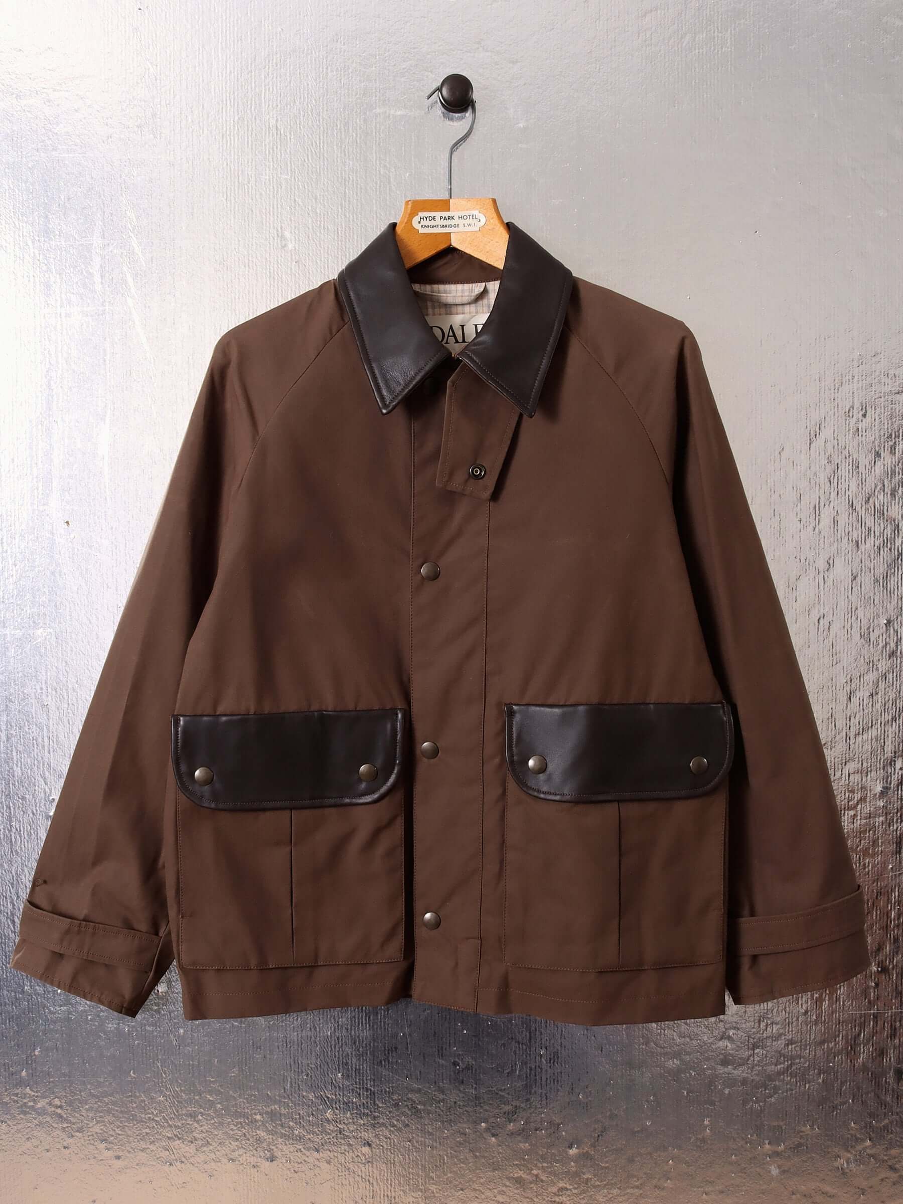 A flat image shows the front of the waxed blouson jacket in dark brown, featuring dark brown leather pocket accents and collar. There are small circular brass button closures and accents on the leather pockets.