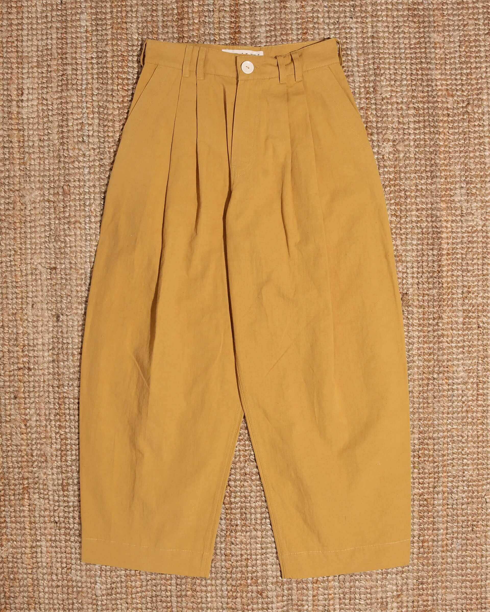 A flat shot of the 'Clarence' Trouser in mustard, showing the relaxed shape of the trouser, the mid-high waist, and double pleated details. 