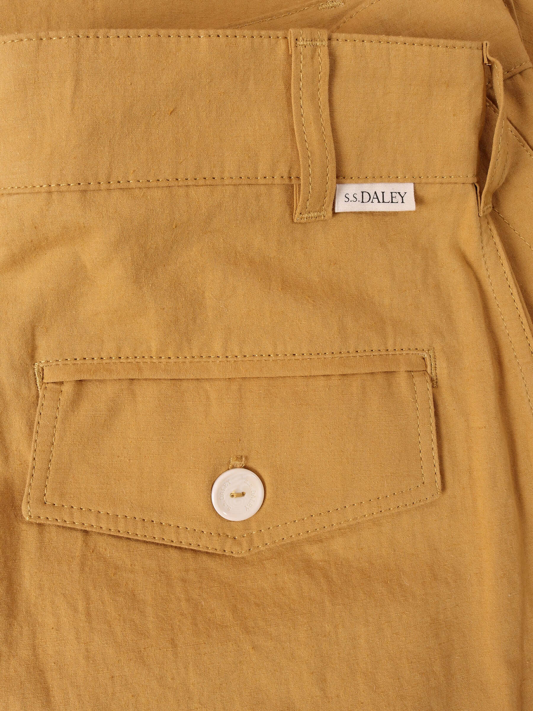 A detail shot of the 'Clarence' Trouser in mustard showing the technical Japanese cotton-blend fabric and the right back pocket with an S.S.DALEY engraved white button, and a small S.S.DALEY logo sitting just next to the belt loop.