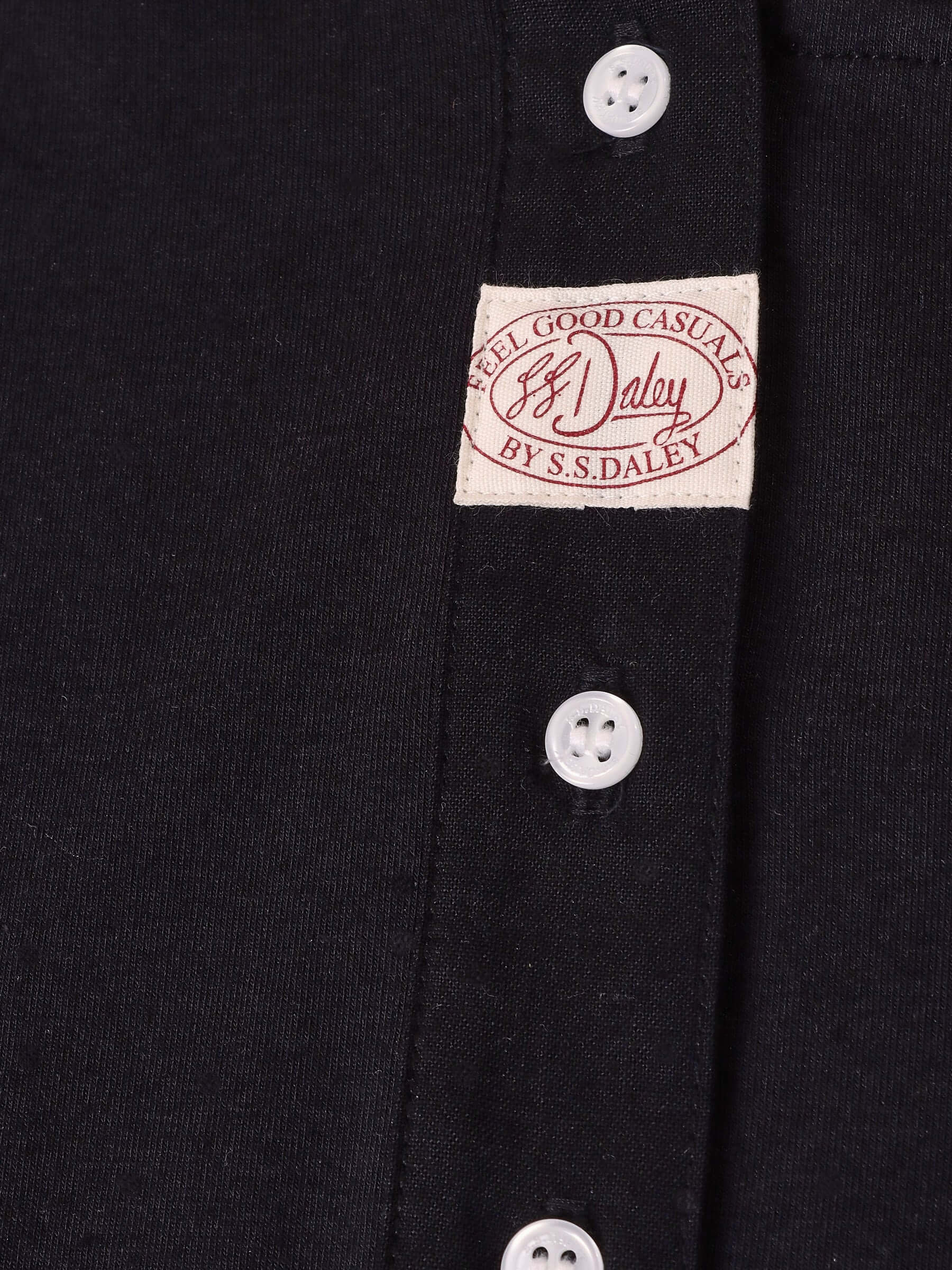 Detail shot of the black 'Aker' Henley's placket with small white buttons and a n S.S.DALEY Feel Good Casuals label.